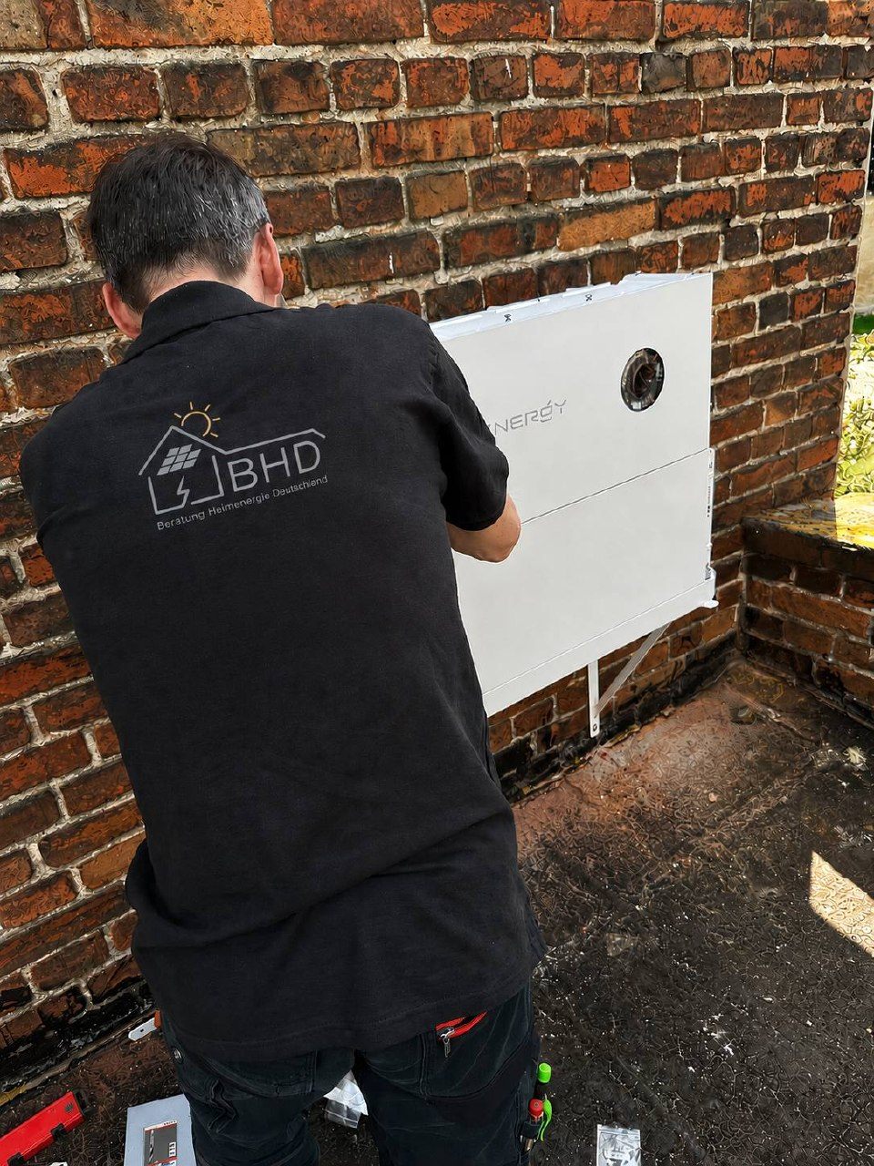 Monteur im BHD-Shirt richtet einen wandmontierten Batteriespeicher an einer Klinkerwand aus