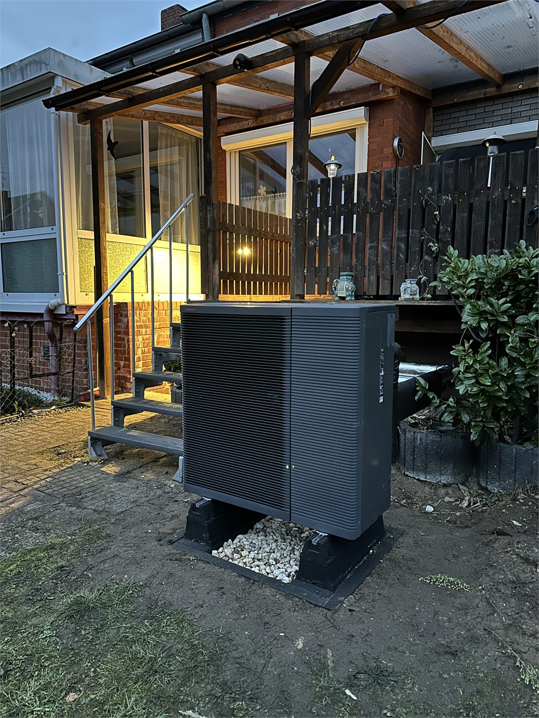 Wärmepumpen-Außengerät direkt an der überdachten Terrasse eines Wohnhauses in der Dämmerung